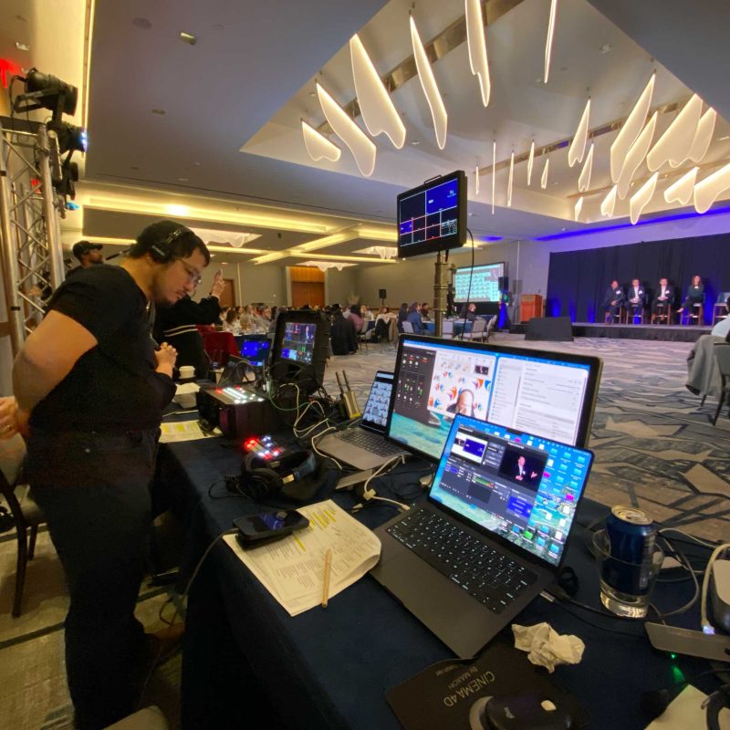 Event video production crew monitors a conference panel and live video feeds from a control station.
