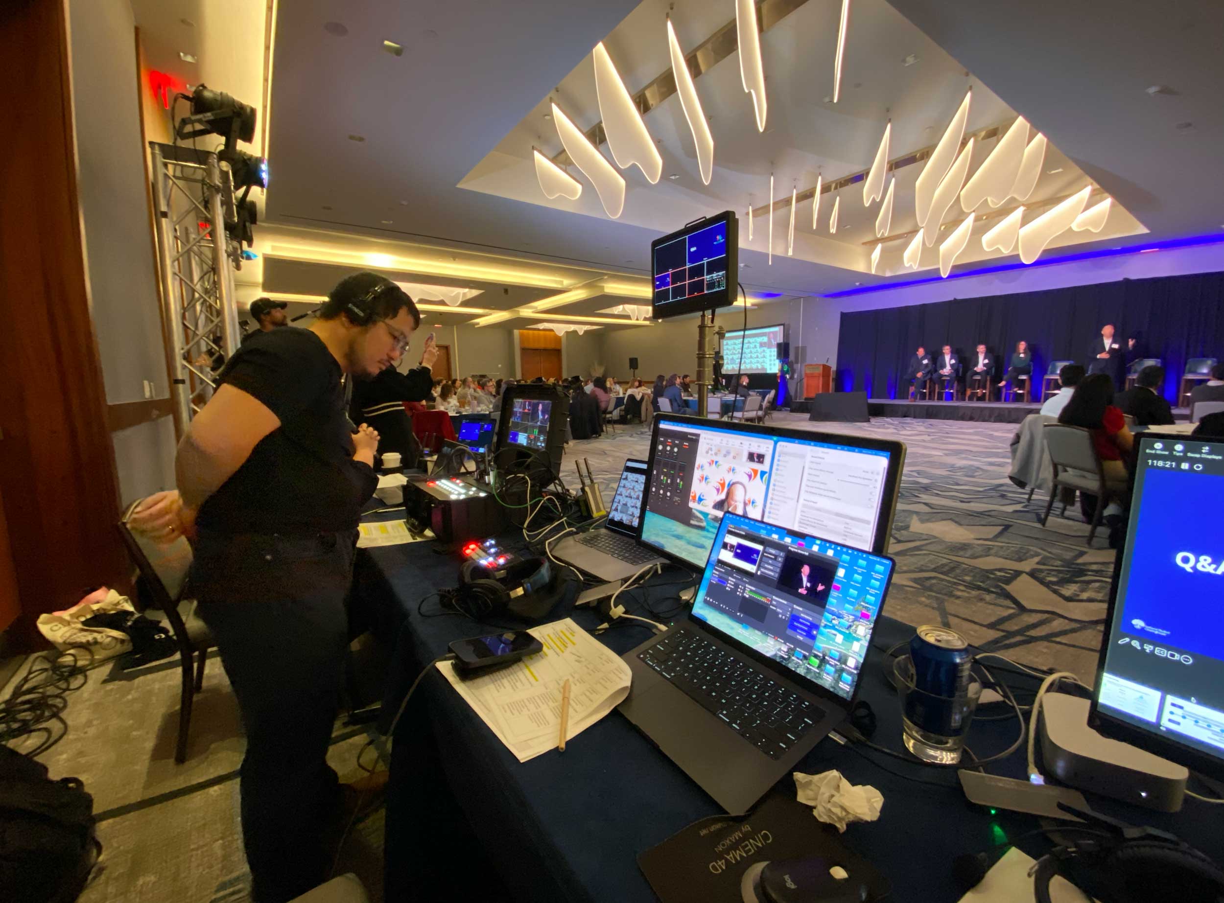 Event video production crew monitors a conference panel and live video feeds from a control station.